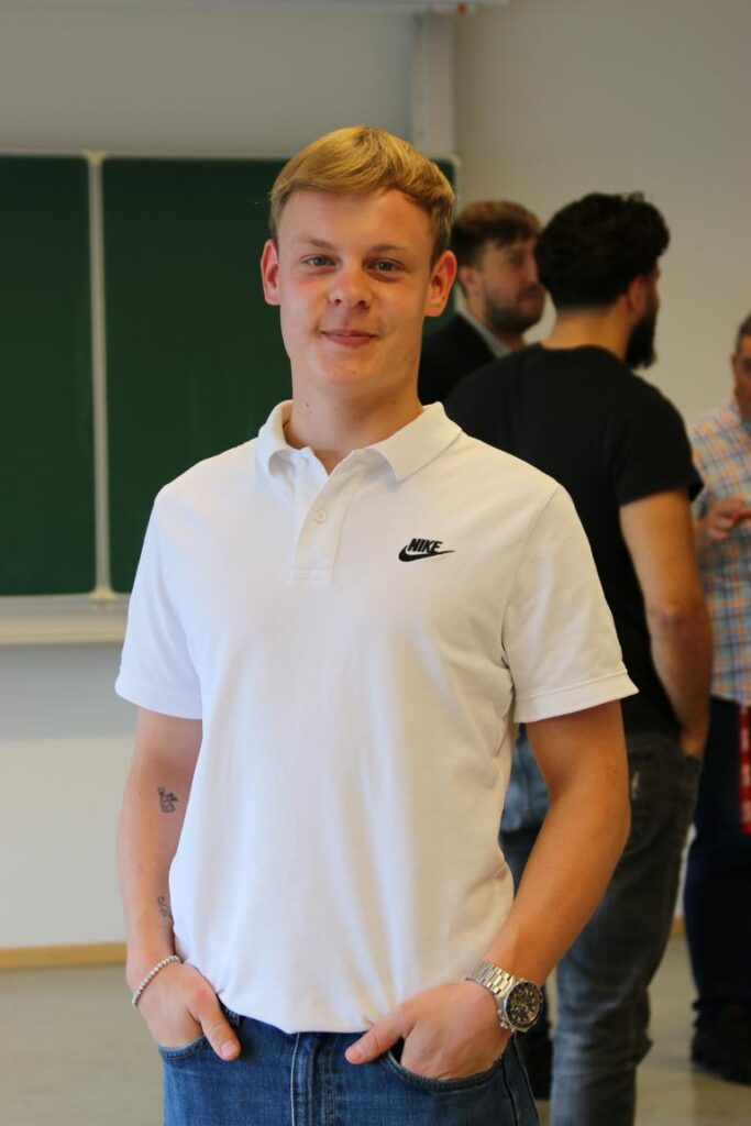 Junger Mann mit blonden Haaren in einem weißen Nike Polohemd steht lächelnd mit beiden Händen in den Hosentaschen, im Hintergrund sind unscharfe Personen und eine Tafel zu sehen.