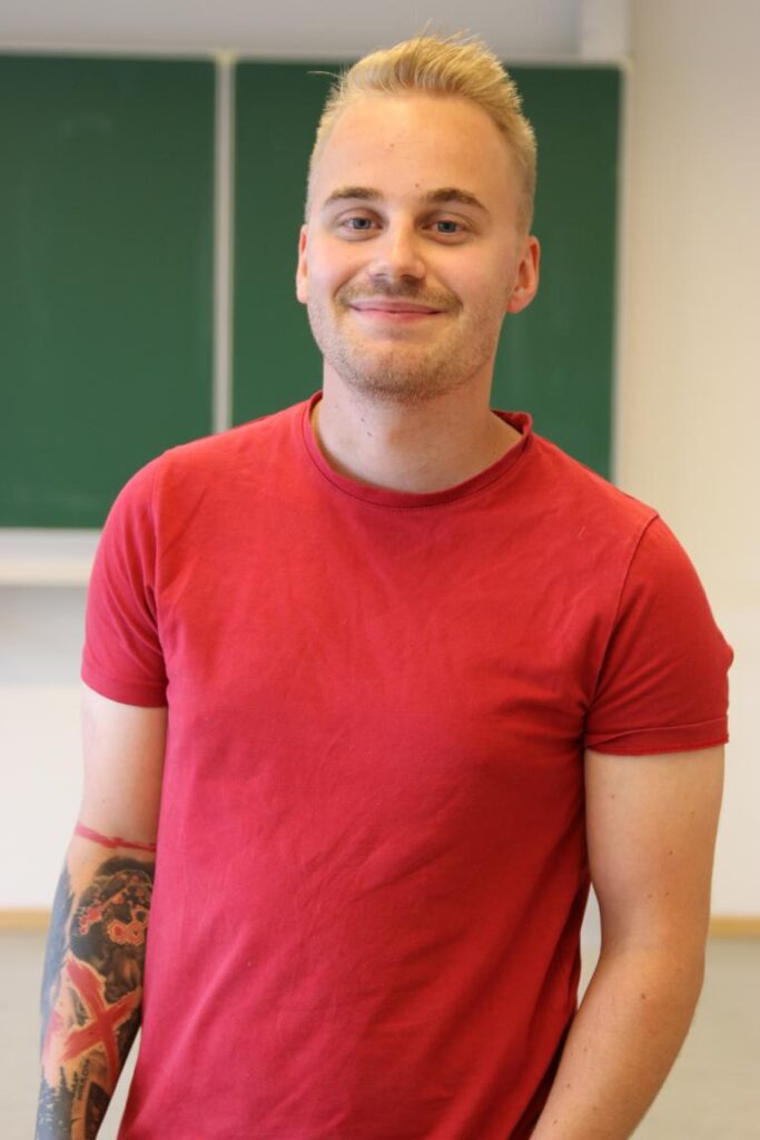 Junger Mann mit blonden Haaren und freundlichem Lächeln, trägt ein rotes T-Shirt und hat einen sichtbaren Tattoo-Verlauf auf dem linken Arm. Im Hintergrund ist eine grüne Tafel zu sehen.