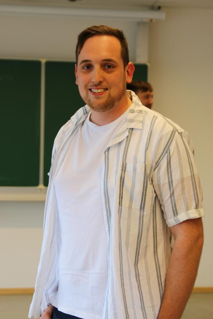 Ein Mann mit kurzen, braunen Haaren und einem leichten Bart steht vor einer grünen Tafel und lächelt, er trägt ein weißes T-Shirt und ein gestreiftes Hemd.