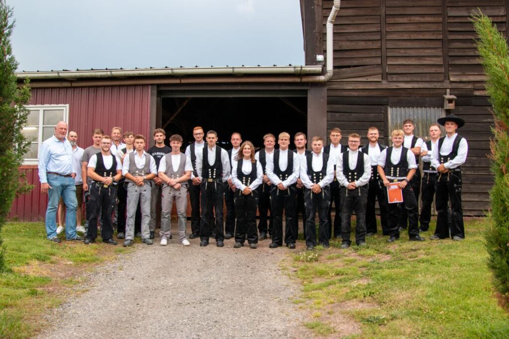 Eine Gruppe von jungen Männern und einer Frau steht in traditioneller Tracht vor einem Holzgebäude. Im Vordergrund sind einige Mitglieder in schwarz-weißer Kleidung mit Westen zu sehen. Im Hintergrund sind weitere Personen, die lässiger gekleidet sind. Der Himmel ist bewölkt.