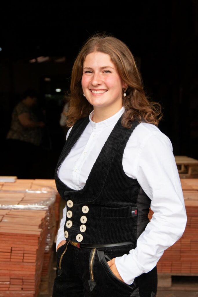 Junge Frau mit schulterlangen, braunen Haaren lächelt in traditioneller Zunftkleidung mit weißem Hemd und schwarzer Weste vor einem Stapel Dachziegel.