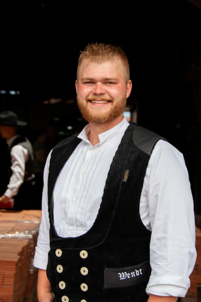Lächelnder junger Mann mit Bart und traditioneller Zimmermannskluft, inklusive weißem Hemd und schwarzer Weste mit auffälligen Knöpfen, steht vor einem dunklen Hintergrund.