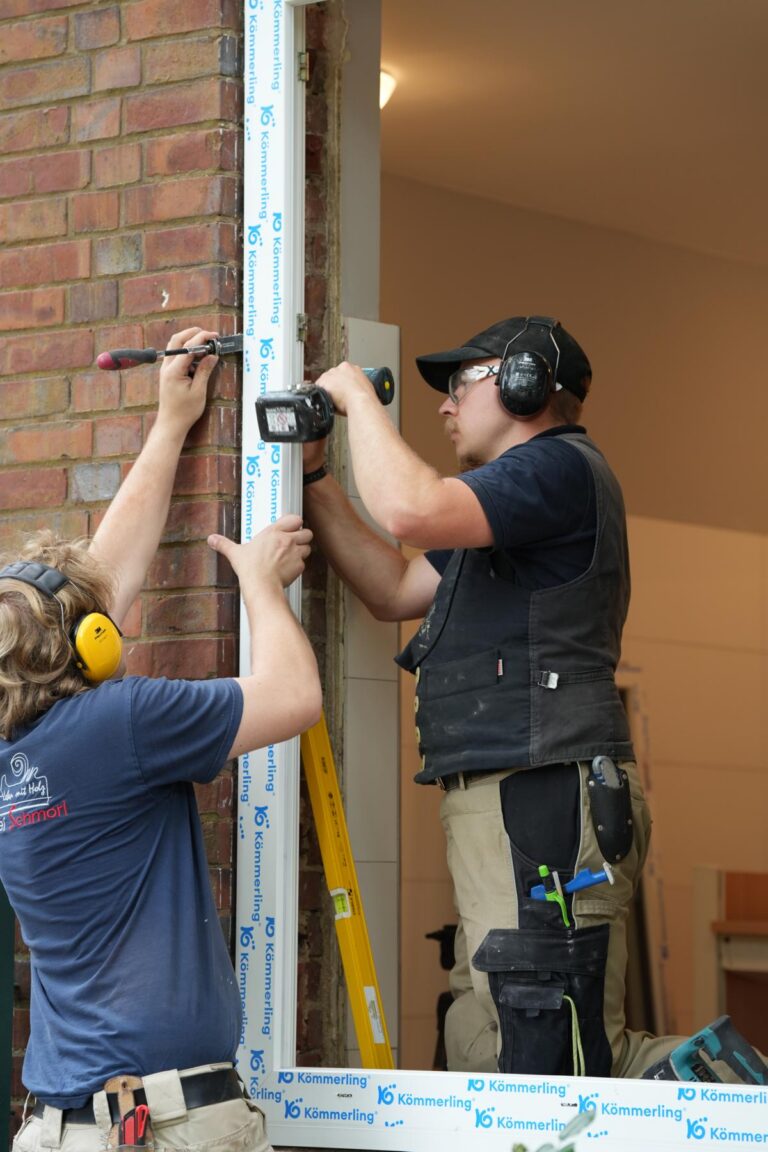 Zwei Handwerker installieren ein Fenster mit einer Werkzeugmaschine, während sie eine Leiter und verschiedene Werkzeuge verwenden. Im Hintergrund ist ein Raum mit neutralen Farben sichtbar. Die Montage beinhaltet eine spezielle Dichtung von Kämmerling.