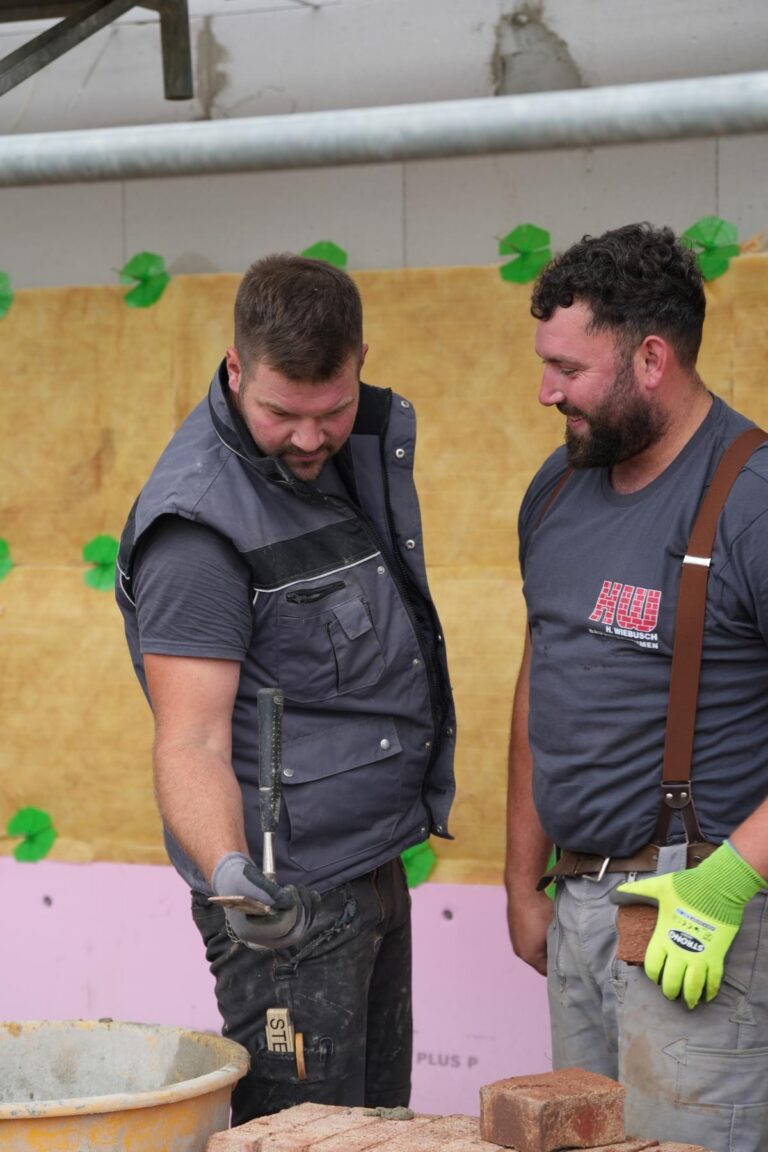 Zwei Bauarbeiter stehen auf einer Baustelle und diskutieren über Mauerarbeiten. Der Mann links hält ein Werkzeug in der Hand, während der andere einen Ziegelstein in seiner Hosentasche hat. Im Hintergrund sind Baumaterialien und eine teilweise verkleidete Wand zu sehen.