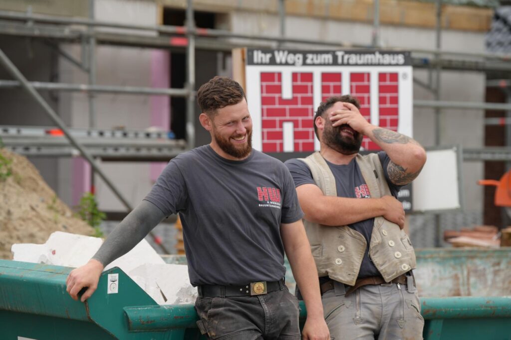 Zwei Bauarbeiter lachen entspannt vor einem Bauprojekt, während sie an einer grünen Abfallcontainer stehen. Im Hintergrund ist ein Schild mit der Aufschrift "Ihr Weg zum Traumhaus" zu sehen.