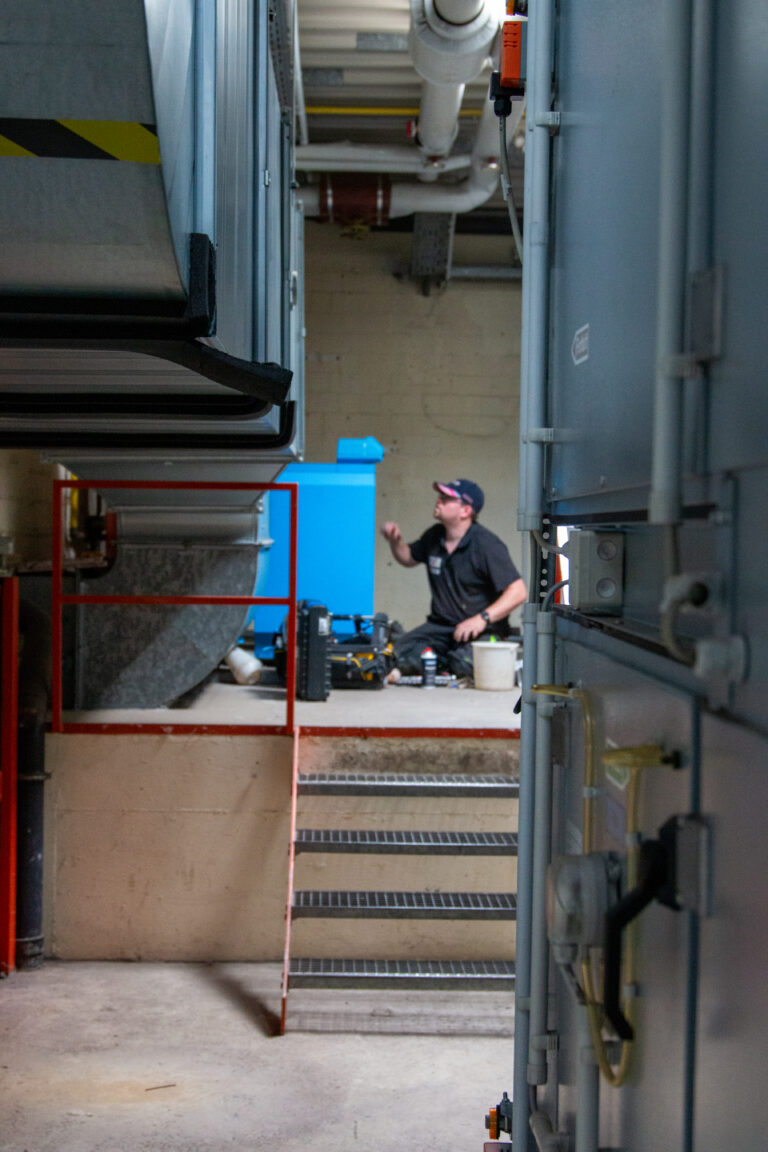 Techniker arbeitet in einem industriellen Bereich, umgeben von Maschinen und Ausrüstung, während er auf einer Treppe sitzt und Werkzeuge nutzt.