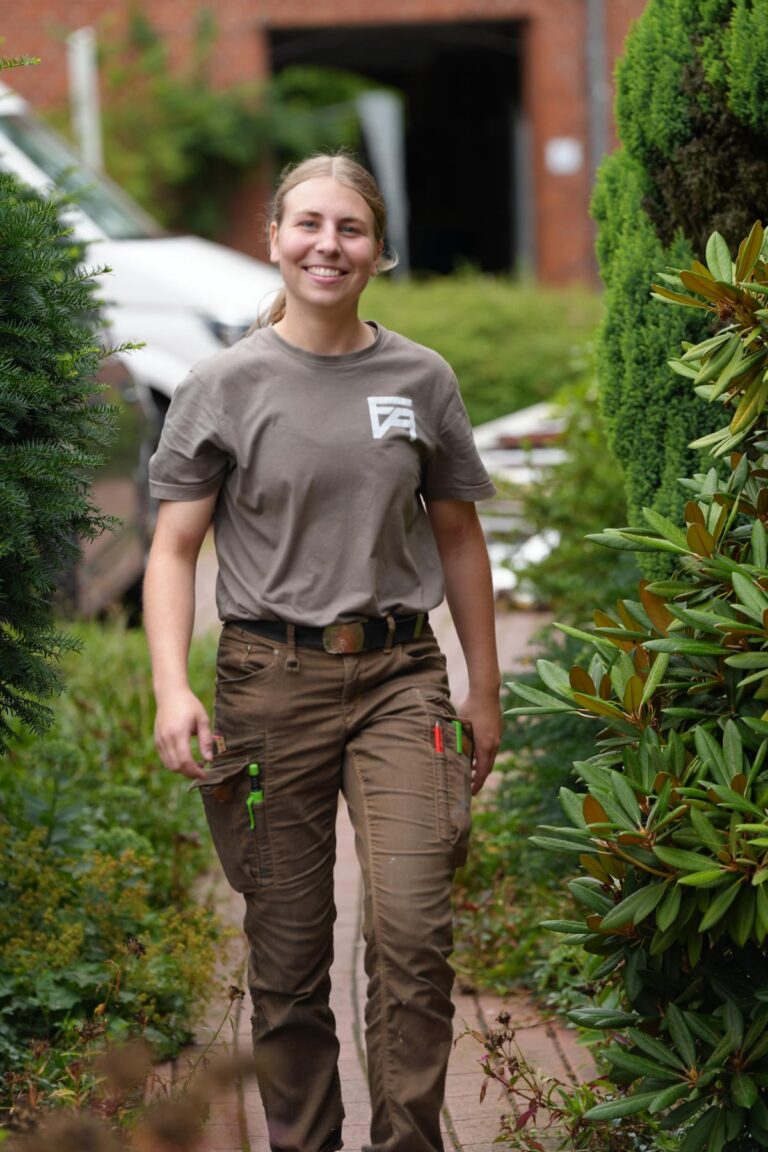 Junge Frau in Arbeitskleidung, die lächelnd einen Gartenweg entlanggeht, umgeben von Pflanzen und Büschen. Sie trägt ein graues T-Shirt und beige Arbeitshosen mit Werkzeugtaschen.