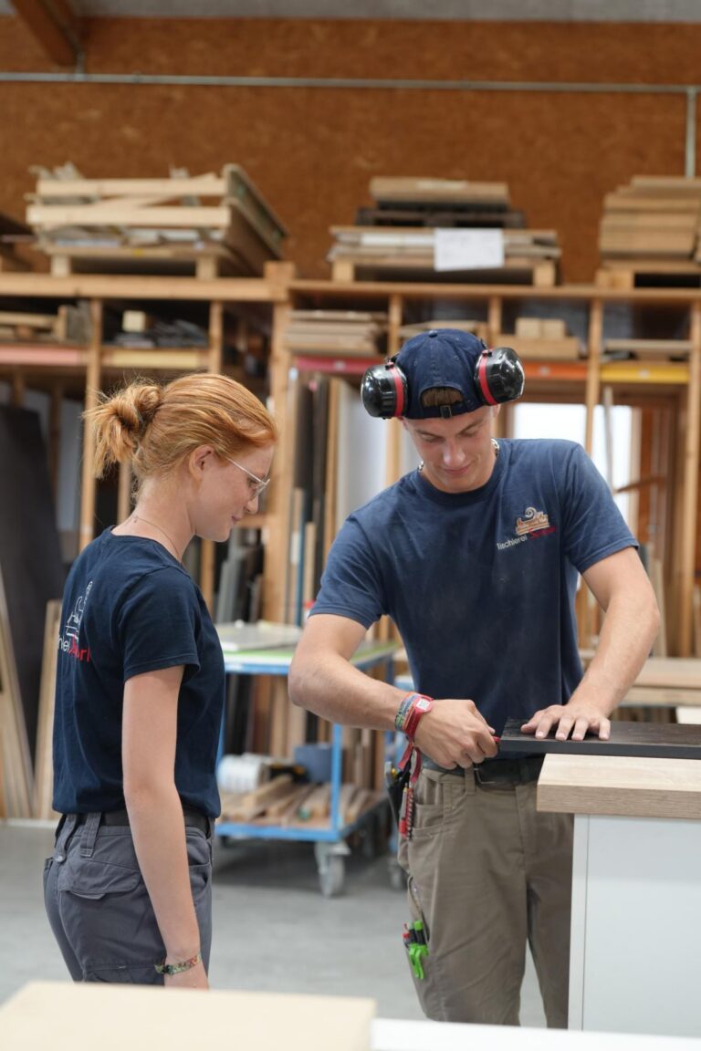 Zwei Auszubildende im Handwerk arbeiten in einer Werkstatt. Die junge Frau mit roten Haaren beobachtet aufmerksam, während der junge Mann mit Gehörschutz ein Stück Holz bearbeitet. Im Hintergrund sind Holzmaterialien und Werkzeuge sichtbar, was die handwerkliche Umgebung unterstreicht.